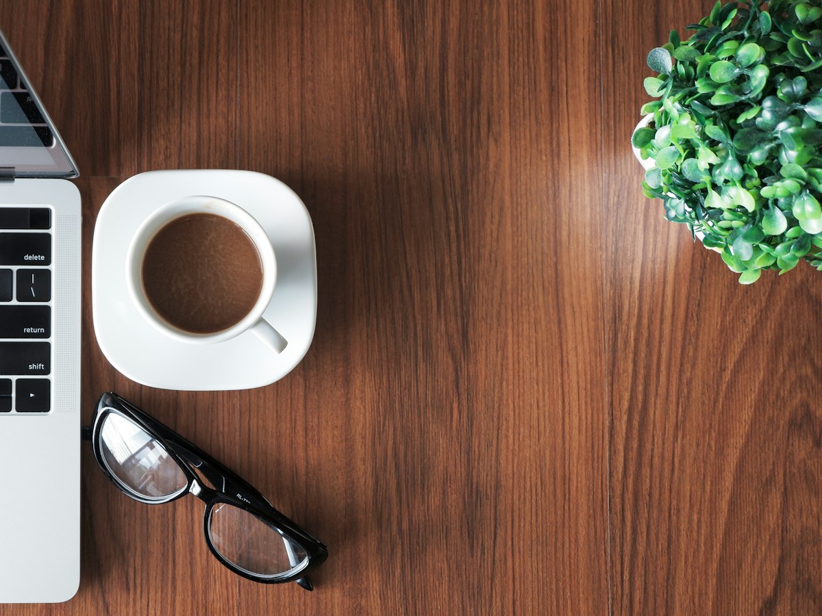 A desk workspace with a laptop, coffee, glasses, and a small plant